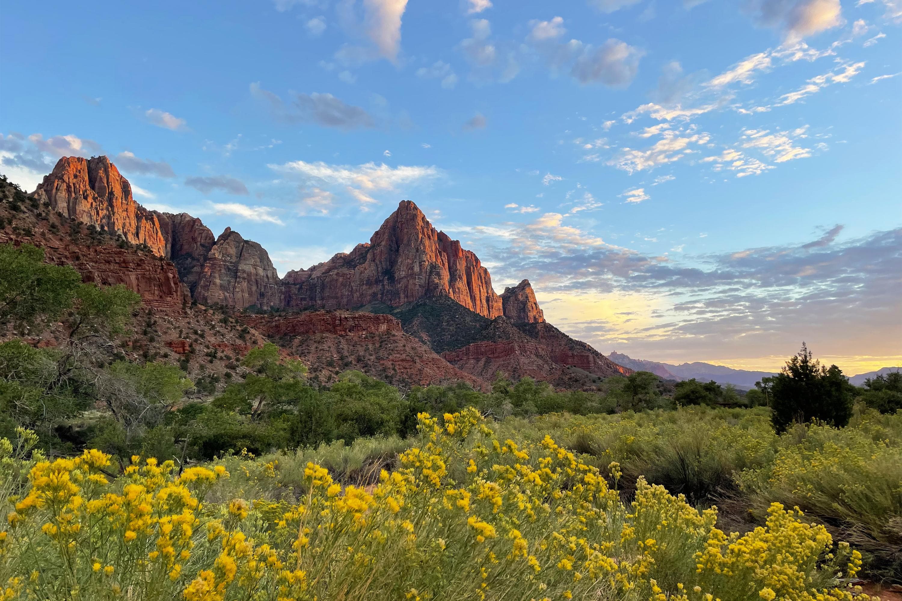 A triangular sandstone mountain rises over green and yellow foliage under a cloudy blue sky.